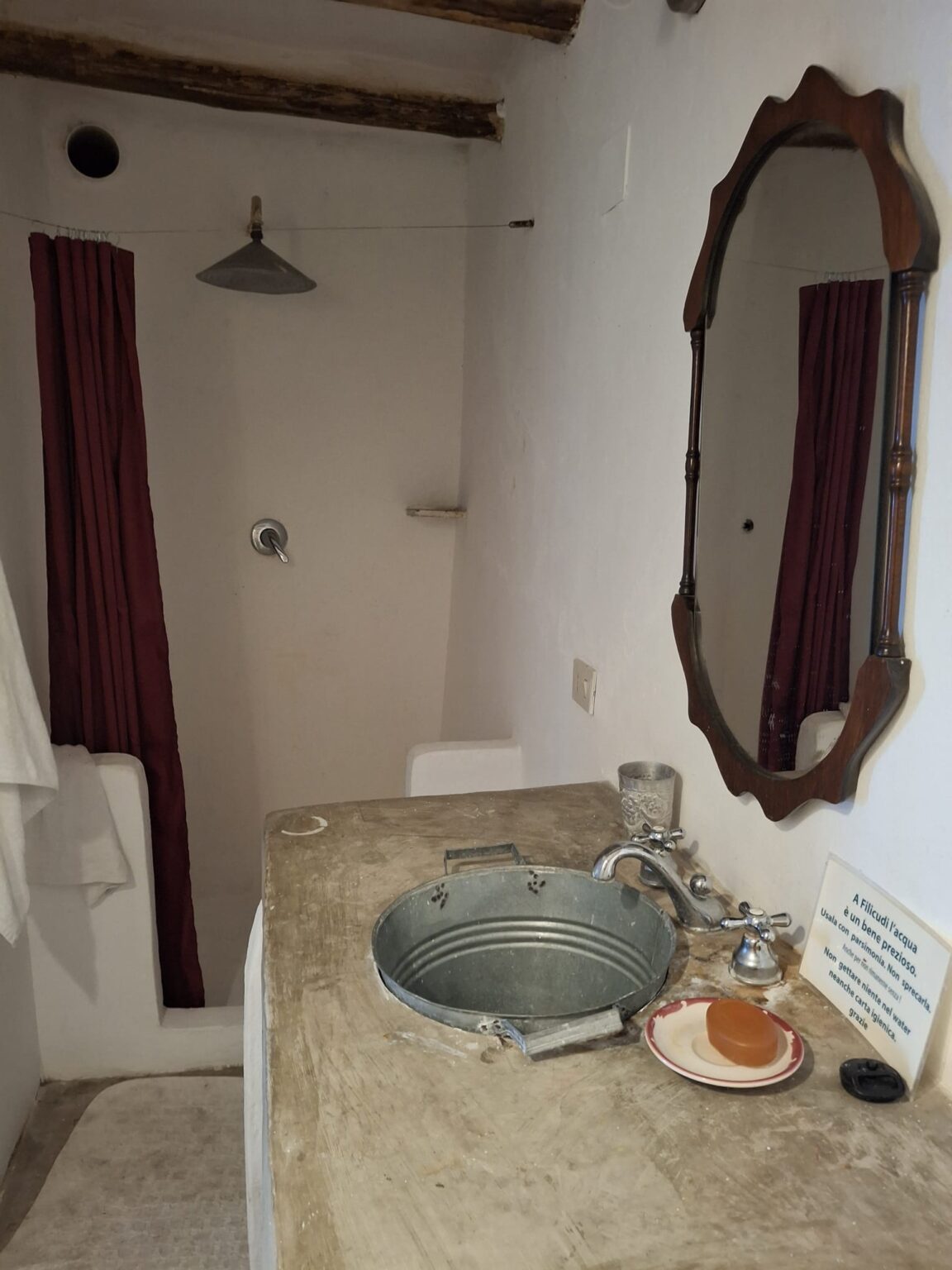 Rustic bathroom featuring a vintage wash basin made from a metal basin, a wooden countertop, and a mirror. The space includes a shower area with maroon curtains and exposed wooden beams. A bar of soap sits on a plate next to the faucet, emphasizing the minimalist design. | Only on SopranoVillas