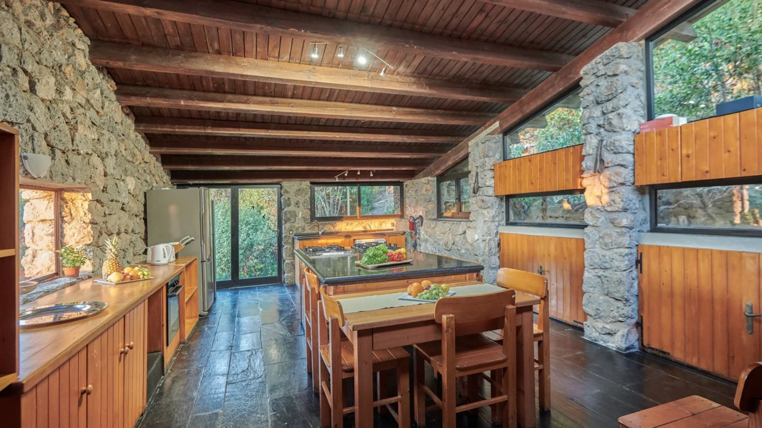Modern kitchen with stone walls, wooden beams, and a large central island. Features include a gas stove, wooden cabinets, and a dining area with chairs. Natural light floods the space through large windows, offering a view of greenery outside. | Only on SopranoVillas