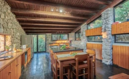 Modern kitchen with stone walls, wooden beams, and a large central island. Features include a gas stove, wooden cabinets, and a dining area with chairs. Natural light floods the space through large windows, offering a view of greenery outside.