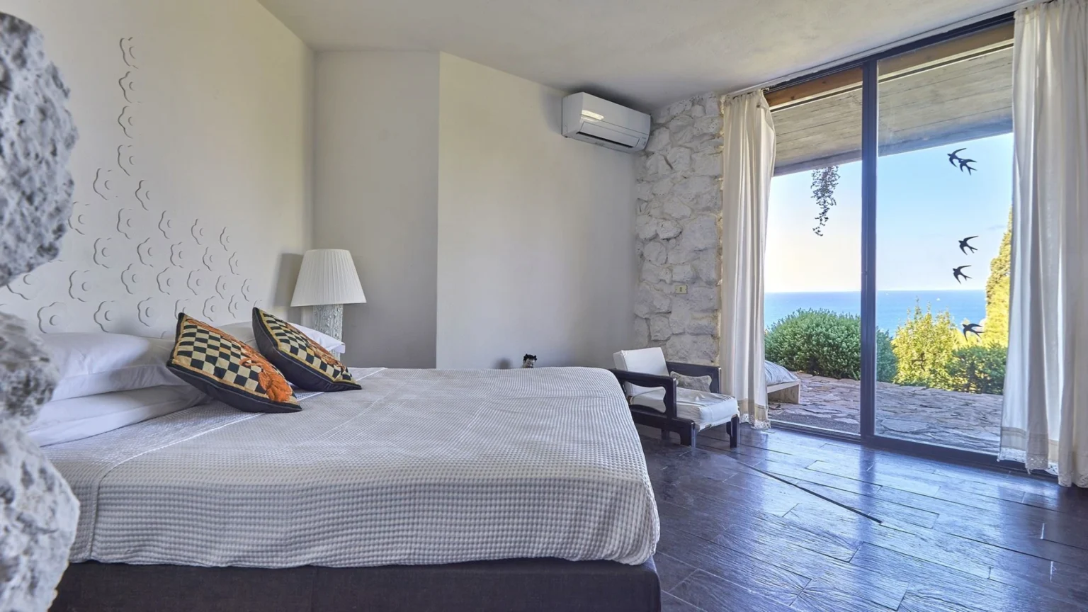 Modern bedroom with a large window showcasing a sea view, featuring a comfortable bed with decorative pillows, a bedside lamp, and minimalist decor including a stone wall and wooden flooring. | Only on SopranoVillas