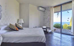 Modern bedroom with a large window showcasing a sea view, featuring a comfortable bed with decorative pillows, a bedside lamp, and minimalist decor including a stone wall and wooden flooring.