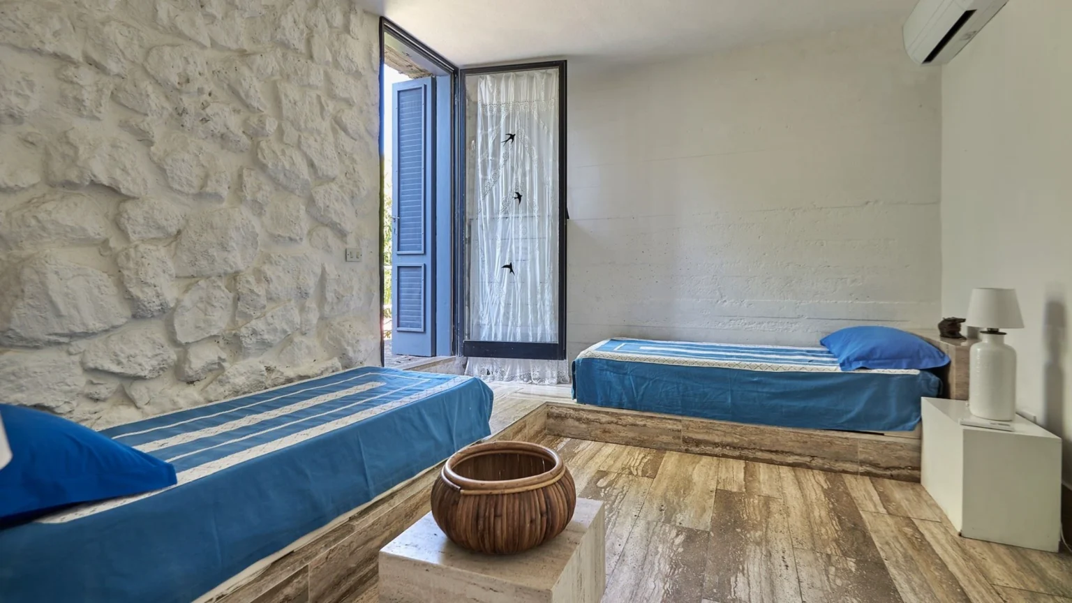 Cozy bedroom featuring two single beds with blue bedding, a stone wall, and a large window opening to a serene outdoor view. The room includes a wooden bowl centerpiece and a bedside lamp, creating a relaxing atmosphere. Ideal for a tranquil getaway. | Only on SopranoVillas