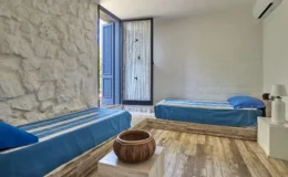 Cozy bedroom featuring two single beds with blue bedding, a stone wall, and a large window opening to a serene outdoor view. The room includes a wooden bowl centerpiece and a bedside lamp, creating a relaxing atmosphere. Ideal for a tranquil getaway.