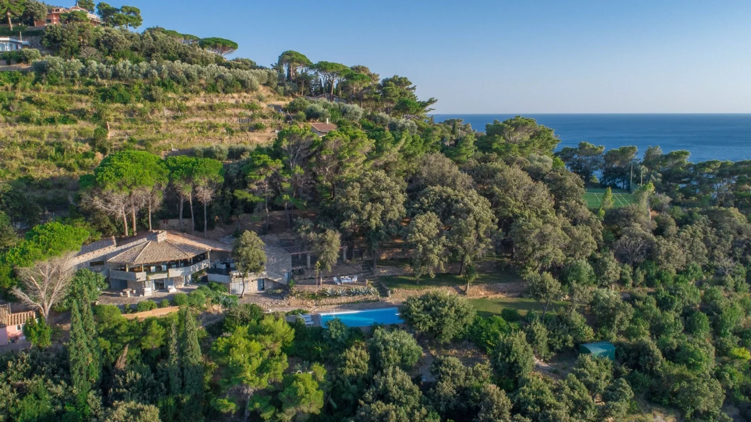 Aerial view of a luxurious hillside villa surrounded by lush greenery, featuring a swimming pool and a tennis court, with the serene ocean in the background. Ideal for vacation rentals and nature retreats. | Only on SopranoVillas