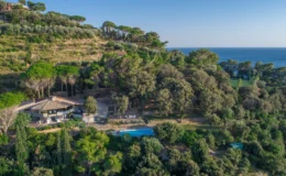 Aerial view of a luxurious hillside villa surrounded by lush greenery, featuring a swimming pool and a tennis court, with the serene ocean in the background. Ideal for vacation rentals and nature retreats.