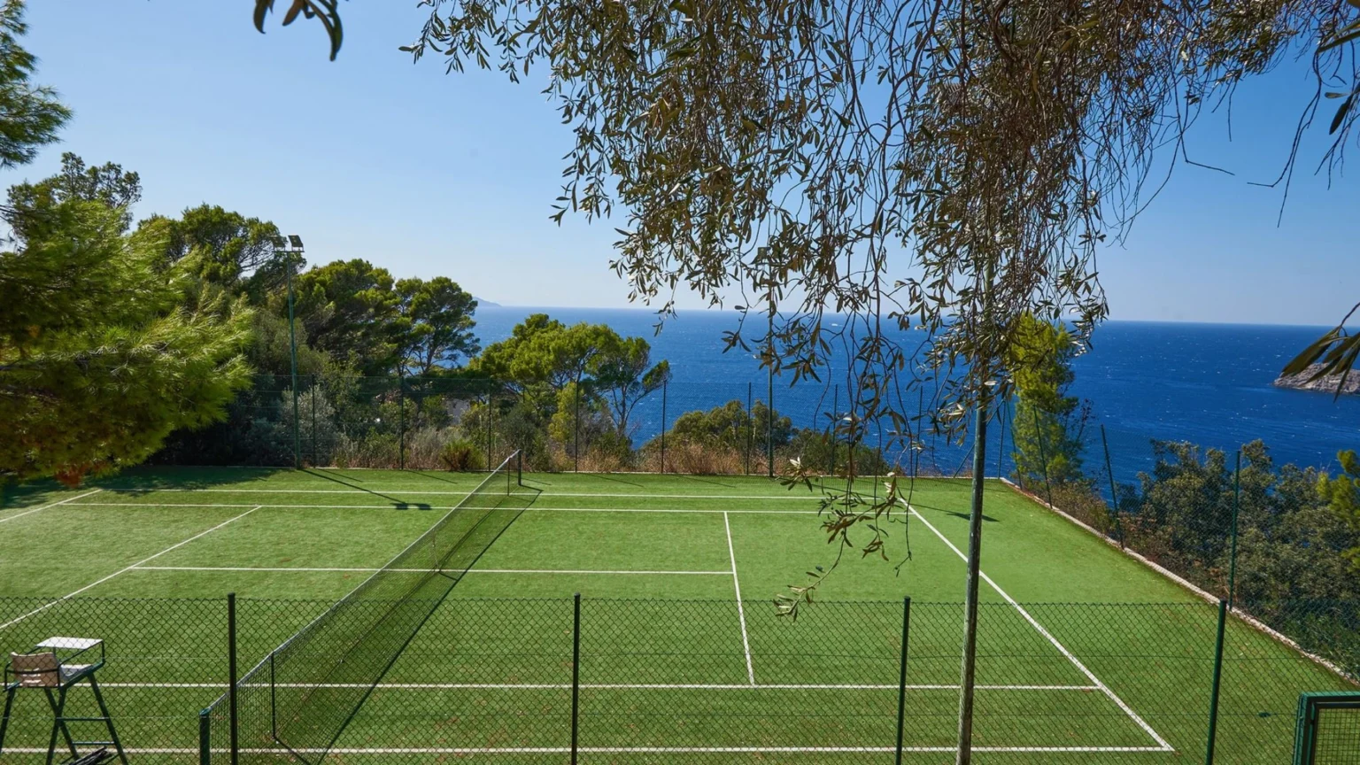 Tennis court with a stunning ocean view, surrounded by lush greenery and tall trees, under a clear blue sky. Ideal for outdoor sports and relaxation. | Only on SopranoVillas