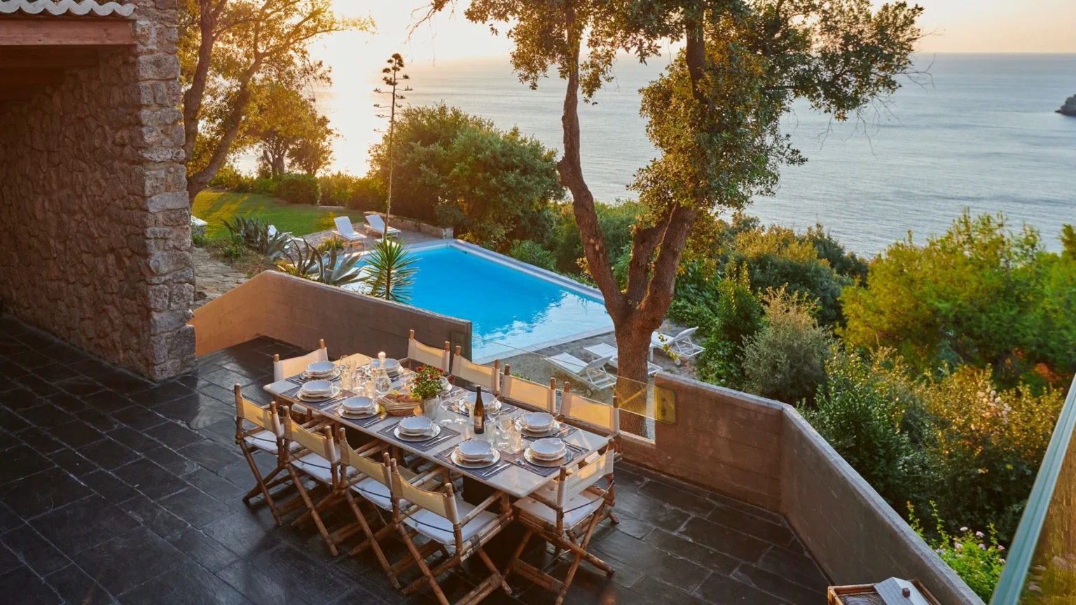 Scenic outdoor dining area with a large table set for a meal, overlooking a swimming pool and the ocean at sunset. Surrounded by lush greenery and stone architecture, this tranquil setting is perfect for entertaining. | Only on SopranoVillas