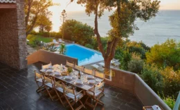 Scenic outdoor dining area with a large table set for a meal, overlooking a swimming pool and the ocean at sunset. Surrounded by lush greenery and stone architecture, this tranquil setting is perfect for entertaining.