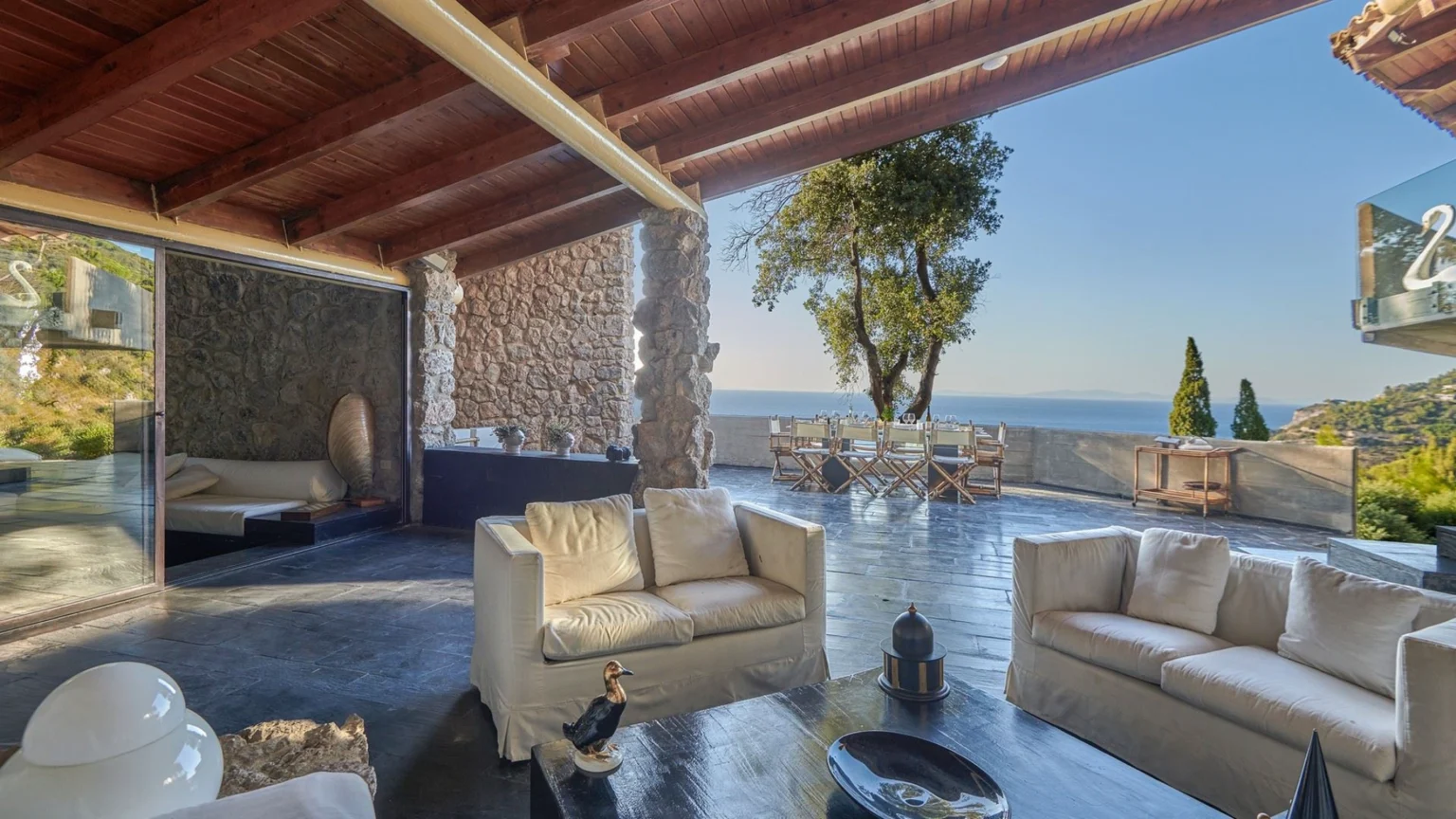 Modern living room with a stunning ocean view, featuring a stone wall, wooden beams, and stylish white sofas. The outdoor dining area is visible through large glass doors, surrounded by greenery, creating a serene and inviting atmosphere. Perfect for relaxation and entertaining. | Only on SopranoVillas