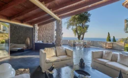 Modern living room with a stunning ocean view, featuring a stone wall, wooden beams, and stylish white sofas. The outdoor dining area is visible through large glass doors, surrounded by greenery, creating a serene and inviting atmosphere. Perfect for relaxation and entertaining.
