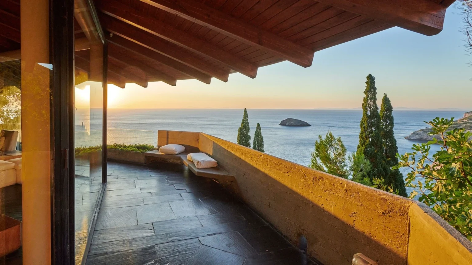 View from a modern balcony showcasing a stunning sunset over the ocean, featuring a sleek stone floor, wooden beams, and plush seating, surrounded by lush greenery and an island in the distance. | Only on SopranoVillas