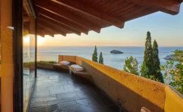 View from a modern balcony showcasing a stunning sunset over the ocean, featuring a sleek stone floor, wooden beams, and plush seating, surrounded by lush greenery and an island in the distance.