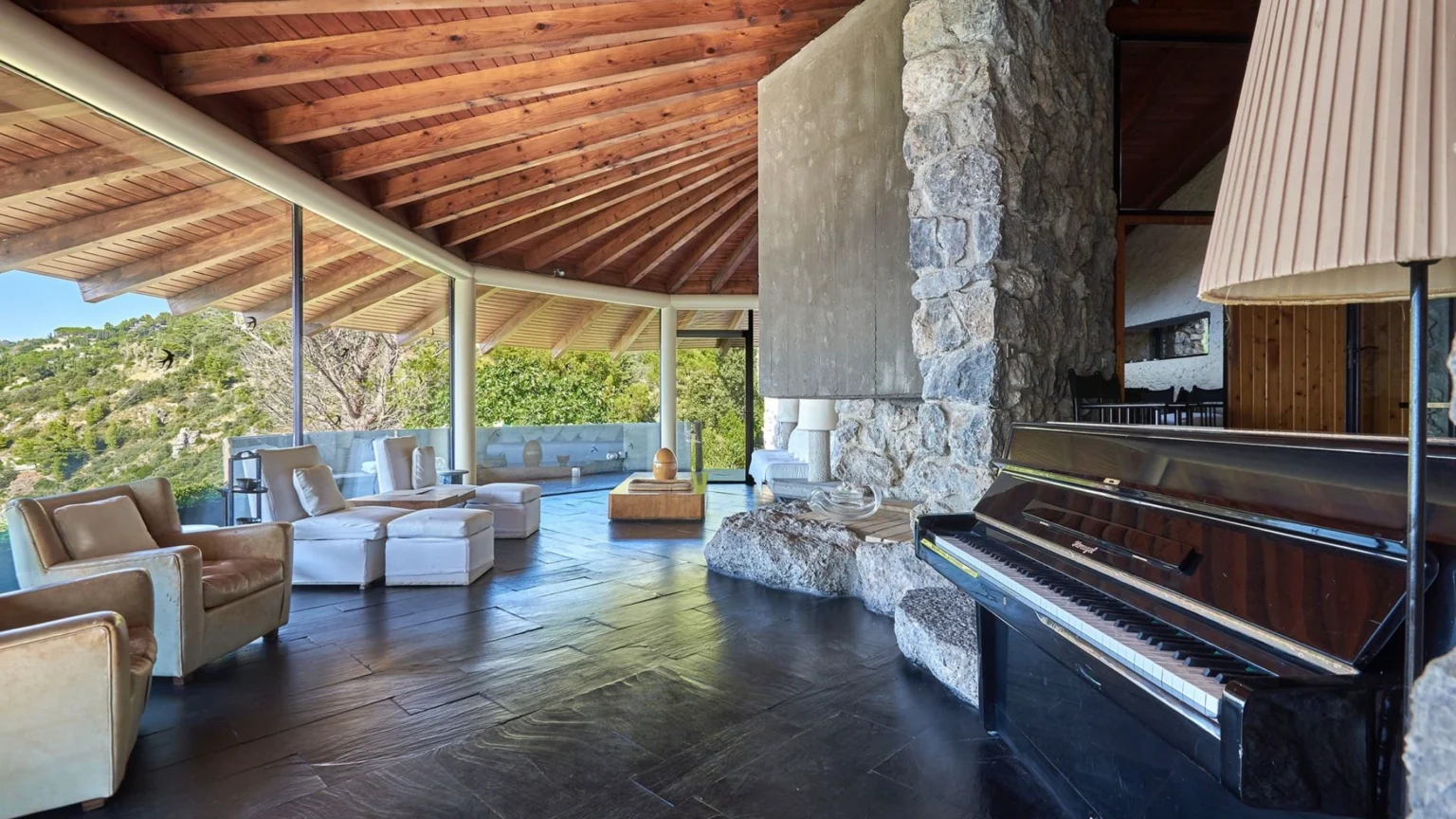 Modern living room with a wooden vaulted ceiling, stone wall, and large glass windows showcasing a scenic view. The space features comfortable seating, a wooden coffee table, and a black piano, creating a stylish and inviting atmosphere. | Only on SopranoVillas
