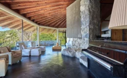 Modern living room with a wooden vaulted ceiling, stone wall, and large glass windows showcasing a scenic view. The space features comfortable seating, a wooden coffee table, and a black piano, creating a stylish and inviting atmosphere.