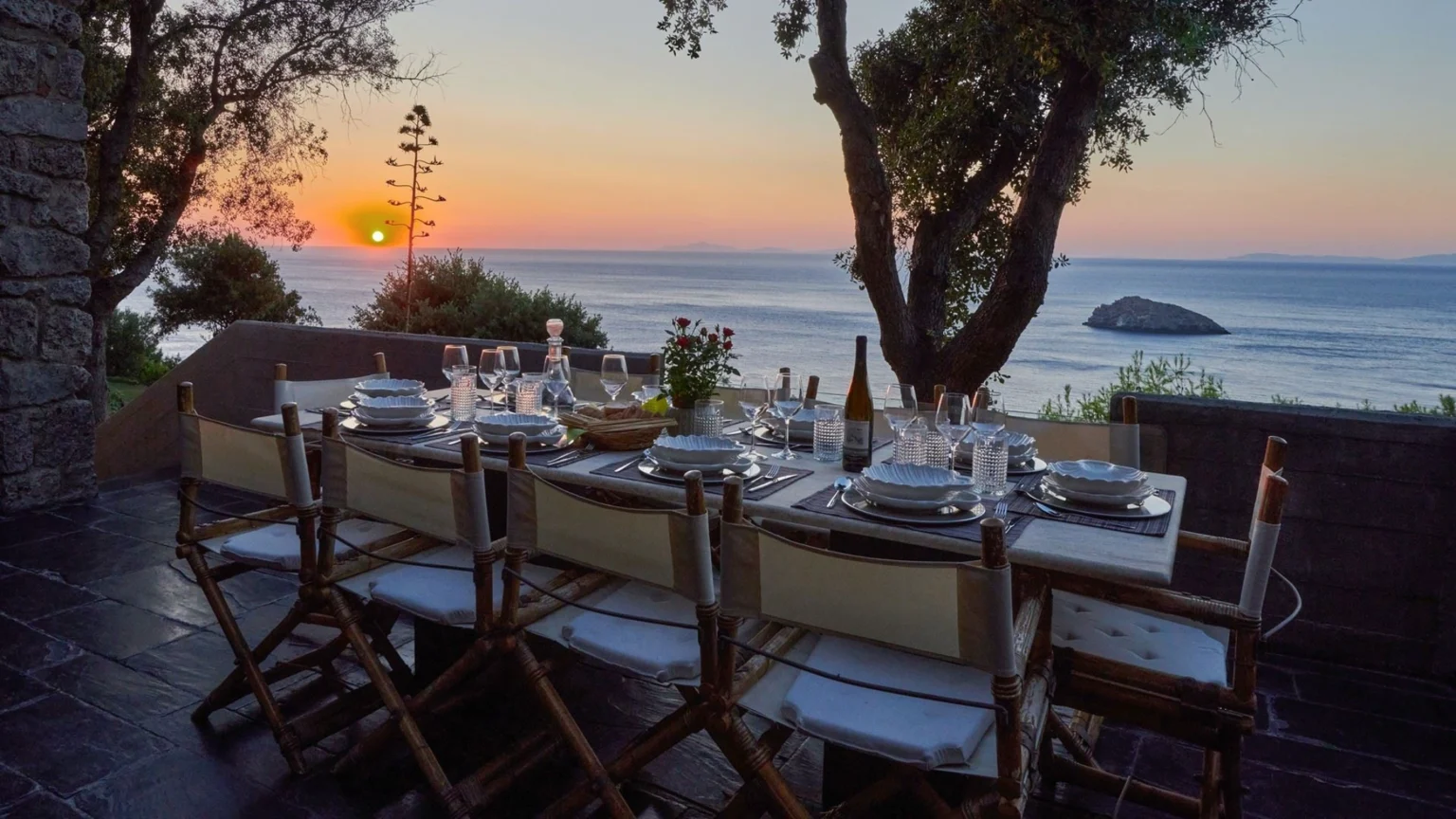 Outdoor dining table set for sunset dinner overlooking the ocean, featuring elegant tableware, wine, and a floral centerpiece, with a scenic view of the setting sun and distant island. | Only on SopranoVillas