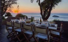 Outdoor dining table set for sunset dinner overlooking the ocean, featuring elegant tableware, wine, and a floral centerpiece, with a scenic view of the setting sun and distant island.
