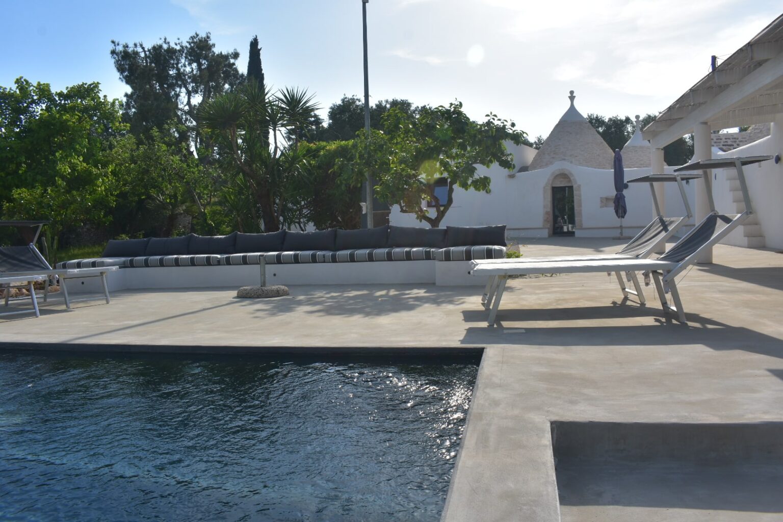 Modern outdoor pool area featuring sleek lounge chairs and a stylish sectional sofa with striped cushions, surrounded by lush greenery and a charming whitewashed building in the background. Ideal for relaxation and leisure. | Only on SopranoVillas
