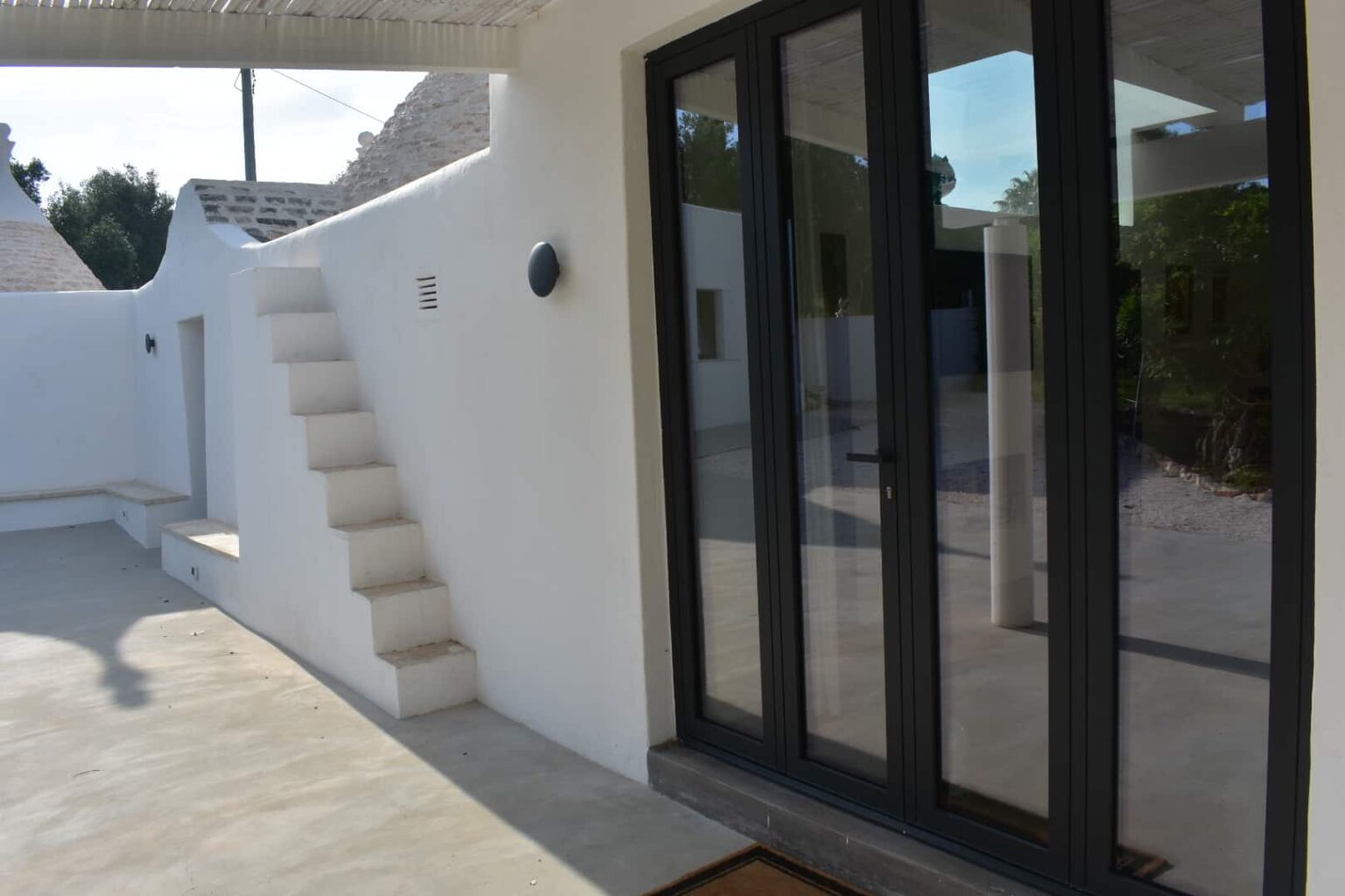 Modern outdoor patio featuring a white stucco wall, sleek black sliding glass doors, and a minimalist staircase. The bright, open space highlights contemporary architectural design and invites natural light. | Only on SopranoVillas