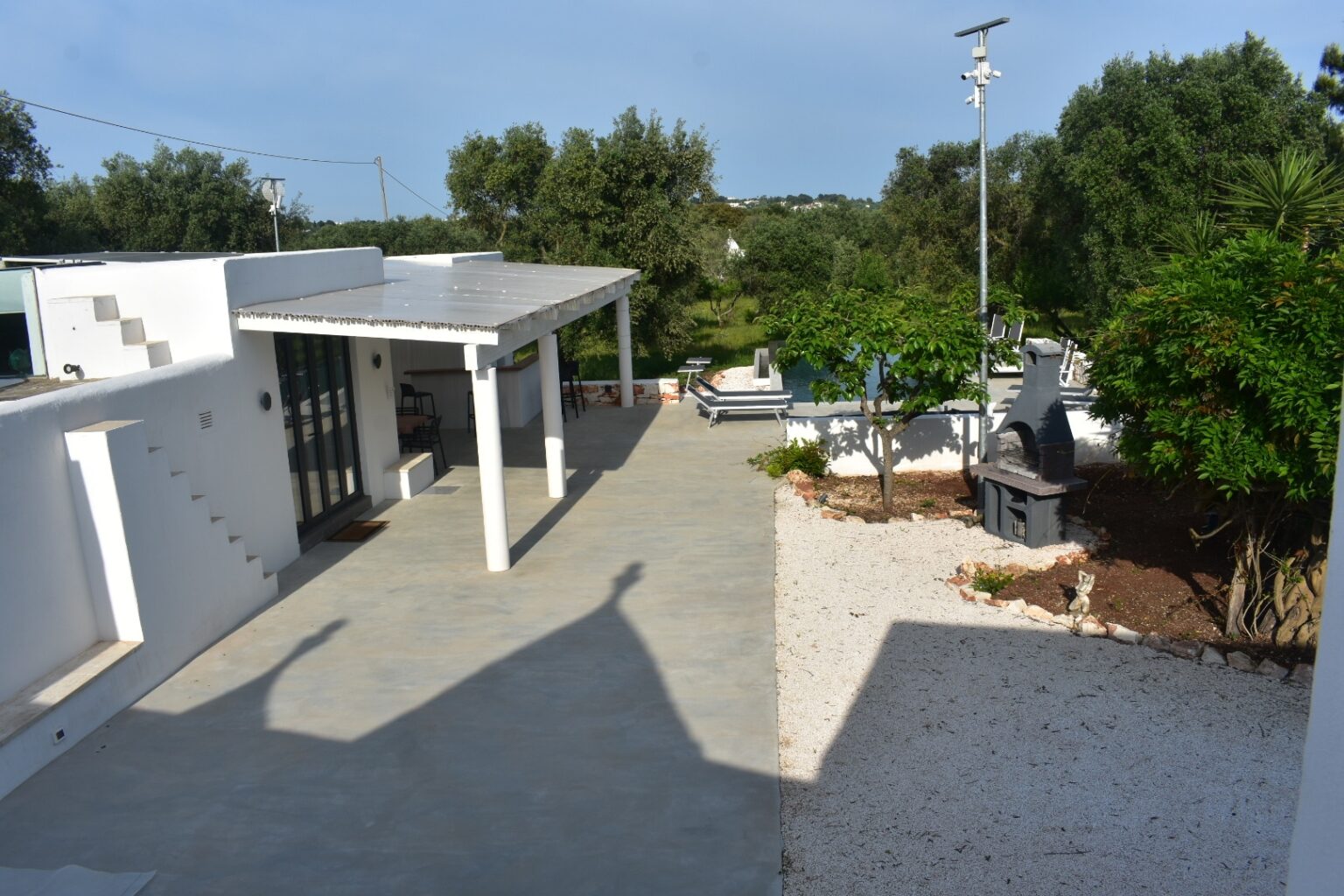 Modern outdoor patio and garden area featuring a spacious concrete pathway, white walls, and lush greenery, ideal for relaxation and entertainment. | Only on SopranoVillas