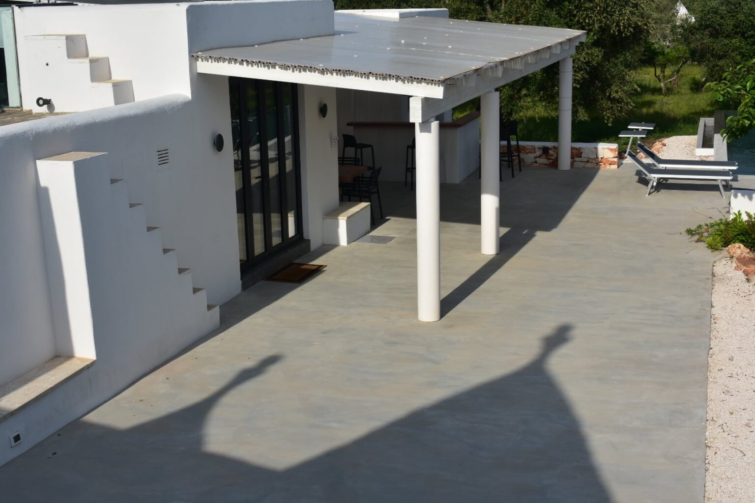 Modern villa exterior with a spacious patio, featuring white walls, a metal roof, and concrete flooring. The image shows steps leading to a glass door, with outdoor furniture including a bar area and loungers visible in a green landscape. | Only on SopranoVillas