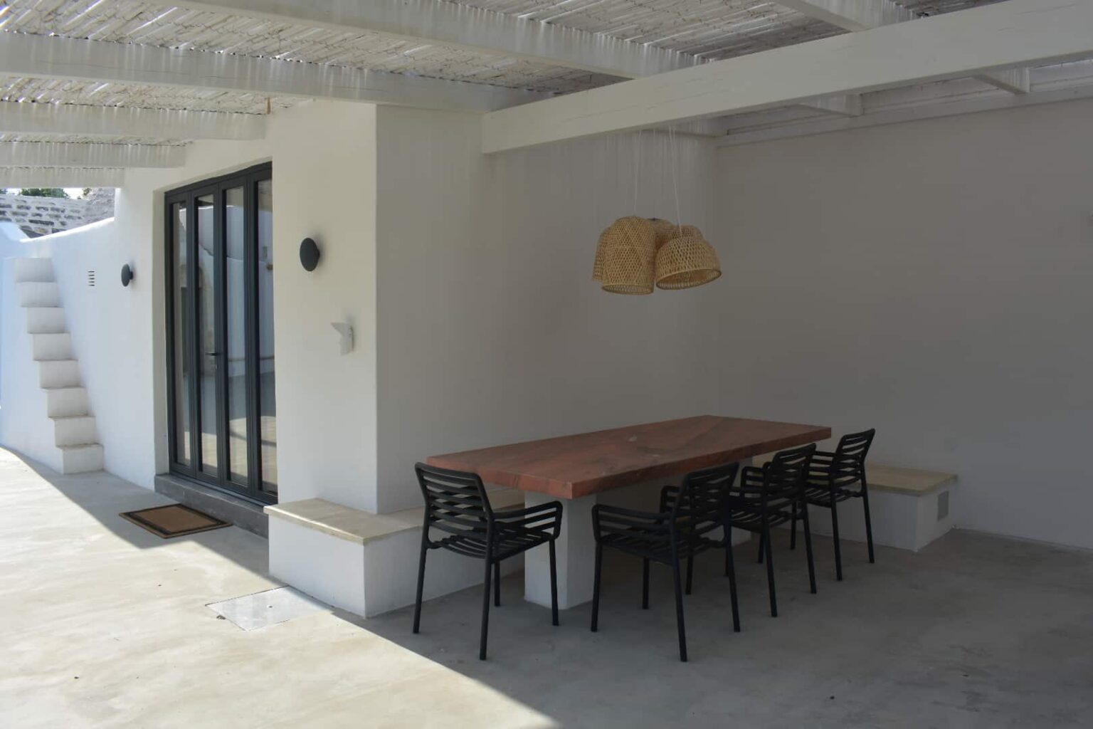 Modern outdoor dining area featuring a wooden table and black chairs, complemented by woven pendant lights and a minimalist white wall design. The setting includes a staircase leading to an upper level, enhancing the contemporary aesthetic. | Only on SopranoVillas