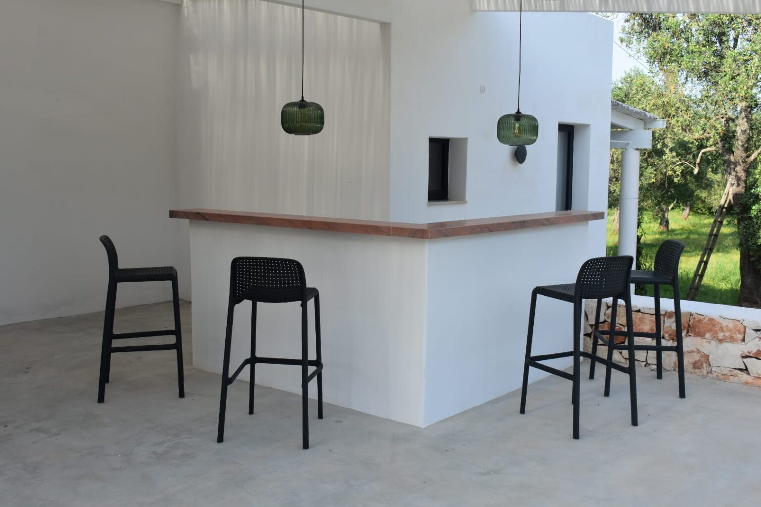 Modern outdoor bar area featuring a sleek white structure with a wooden countertop, complemented by four black high stools and stylish green pendant lights. Lush greenery surrounds the setting, creating a tranquil atmosphere. Ideal for entertaining or relaxing. | Only on SopranoVillas