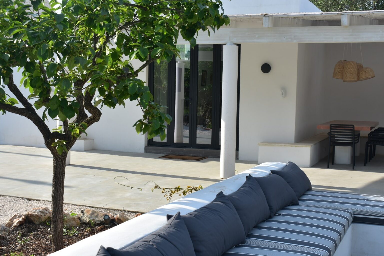 Modern outdoor living space featuring a comfortable sofa with gray cushions, a lush green tree, and a contemporary home with large glass doors. This serene setting emphasizes relaxation and stylish design, perfect for entertaining or enjoying nature. | Only on SopranoVillas