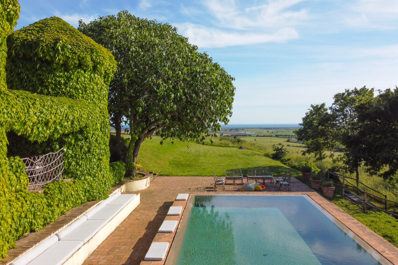 Scenic view of a luxurious villa with a swimming pool surrounded by lush greenery and expansive countryside, featuring a large tree and distant ocean views. Ideal for relaxation and outdoor living. | Only on SopranoVillas