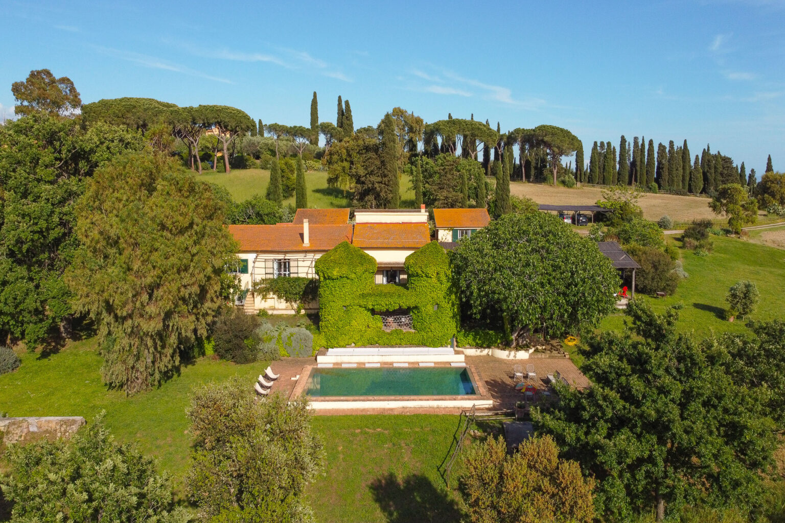 Aerial view of a charming countryside villa surrounded by lush greenery, featuring a swimming pool and landscaped gardens, with tall cypress trees in the background under a clear blue sky. | Only on SopranoVillas