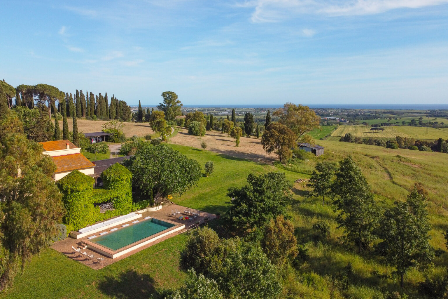 Aerial view of a picturesque countryside villa featuring a swimming pool, surrounded by lush greenery and rolling hills, with a clear blue sky and distant ocean view. | Only on SopranoVillas