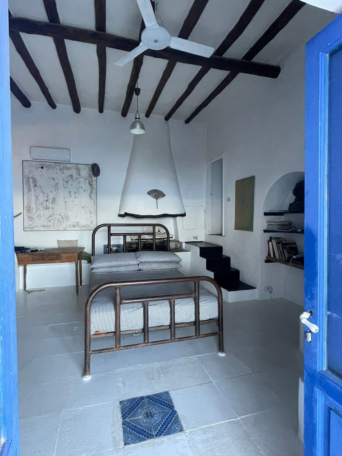 Cozy bedroom interior with wooden beams, a ceiling fan, and a vintage metal bed frame. The room features whitewashed walls, a striped bedding set, a wooden desk, and a staircase leading to an upper level. A decorative tile adds character to the floor, while a blue door frames the entrance. | Only on SopranoVillas