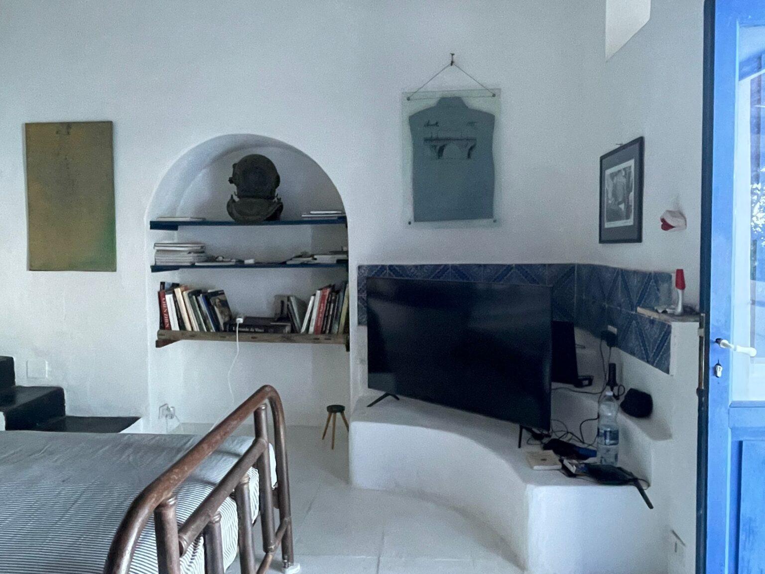 Cozy interior of a modern living space featuring a bed with a metal frame, a TV mounted on a white wall, and a blue door leading outside. The room includes shelves filled with books, decorative art pieces, and a unique sculpture, all set against a backdrop of white walls and blue tiled accents. | Only on SopranoVillas