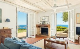 Cozy living room with ocean view, featuring a comfortable blue sofa, wooden coffee table, and a fireplace. Large open doors lead to a scenic outdoor area with greenery and coastal landscape. Bright, airy atmosphere with natural light and ceiling fan.