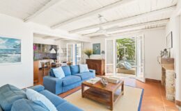 Bright and airy living room featuring blue sofas, a wooden coffee table, and an open layout that connects to a sunny outdoor space. The room has a coastal decor theme with artwork and large windows allowing natural light to fill the space.