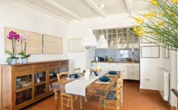 Bright and airy kitchen dining area featuring a wooden table set for a meal, surrounded by chairs. The space includes a stylish wooden sideboard, vibrant orchid plants, and modern pendant lighting, complemented by blue tiled backsplash in the kitchen.
