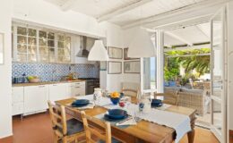 Bright and airy dining area featuring a wooden table set for a meal, surrounded by light-colored chairs, with a view of an outdoor patio through open doors. The kitchen in the background showcases blue tiled backsplash and modern appliances, creating a warm and inviting atmosphere.