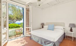 Bright and airy bedroom with a large bed, featuring a light blue accent pillow, white walls, and terracotta flooring. Open French doors lead to a scenic outdoor view of greenery and a sea landscape. Two bedside lamps add a cozy touch. Ideal for a relaxing getaway.