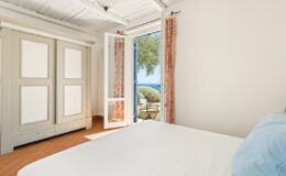 Cozy bedroom with a double bed, featuring a light color scheme, a wardrobe, and large open doors leading to a scenic outdoor view of the sea and greenery.