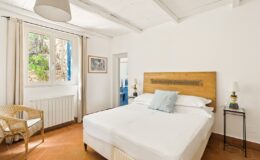 Bright and inviting bedroom featuring a double bed with a wooden headboard, a cozy wicker chair, and a small bedside table with a lamp. Natural light streams in through a large window adorned with light curtains, revealing a view of lush greenery outside. The room showcases terracotta flooring and a minimalist design, creating a serene atmosphere.