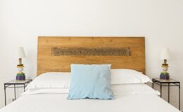 Modern bedroom interior featuring a wooden headboard, a light blue decorative pillow, and stylish bedside lamps on black metal tables. The minimalist design emphasizes a clean and inviting atmosphere.