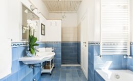 Bright and modern bathroom featuring blue tiled walls, a white sink with a mirror, a bathtub, and stylish decor including a potted orchid. Natural light floods the space through window blinds, enhancing the clean and inviting atmosphere.
