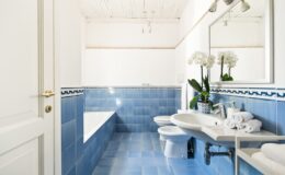 Modern bathroom interior featuring blue tiled walls and floor, a white bathtub, and a sleek sink with towels and decorative plants. Bright and airy design with natural light.