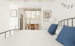 Bright and airy bedroom featuring a minimalist design with a white bed, blue accent pillows, and natural light streaming through a large window. A small wooden desk and artwork adorn the space, creating a cozy and inviting atmosphere. Ideal for relaxation and comfort.