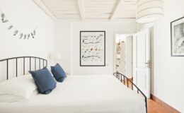 Bright and airy bedroom featuring a double bed with blue accent pillows, white walls, and wooden flooring. The room includes decorative artwork, a modern light fixture, and an open doorway leading to another space, creating a welcoming atmosphere.