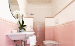 Modern bathroom interior featuring pink tiled walls, a round mirror, a white sink with a towel, and a toilet. Decorative potted orchids add a touch of elegance to the space.