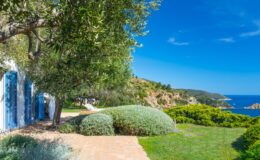 Coastal view of a serene villa with blue shutters, surrounded by lush greenery and overlooking a clear blue sea under a bright sky. Ideal for vacation rentals or holiday retreats.