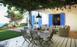 Cozy outdoor dining area with a white table set for a meal, surrounded by wicker chairs under a vine-covered pergola, overlooking a lush garden and sea view. Bright blue shutters complement the modern architectural style of the home.