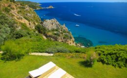 Aerial view of a coastal landscape featuring lush greenery, rocky cliffs, and a vibrant blue sea, with sun loungers positioned on a grassy area overlooking the water.