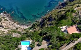 Aerial view of a coastal property featuring a swimming pool, lush greenery, and rocky shoreline beside clear blue waters. Ideal for vacation rentals or seaside retreats.