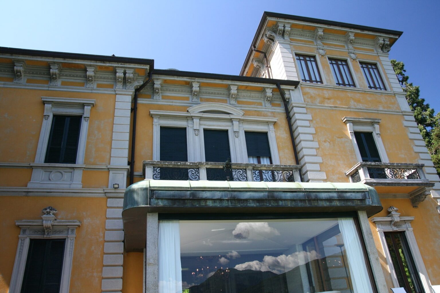 Historic villa with yellow exterior, featuring large windows and a balcony, set against a clear blue sky. Ideal for showcasing architectural beauty and heritage. | Only on SopranoVillas